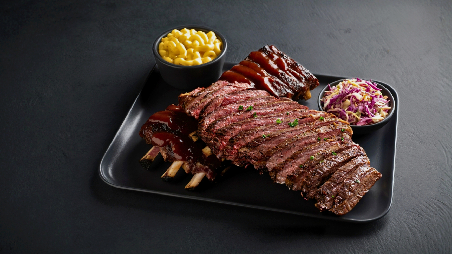 Sliced smoked brisket and baby back ribs on a black tray with mac and cheese and coleslaw